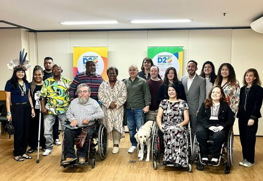 Foto de um grupo de cerca de 10 pessoas ladeando a Ministra dos Direitos Humanos e da Cidadania, Macaé Evaristo, e a Secretária Nacional dos Direitos da Pessoa com Deficiência do MDHC, Anna Paula Feminella.