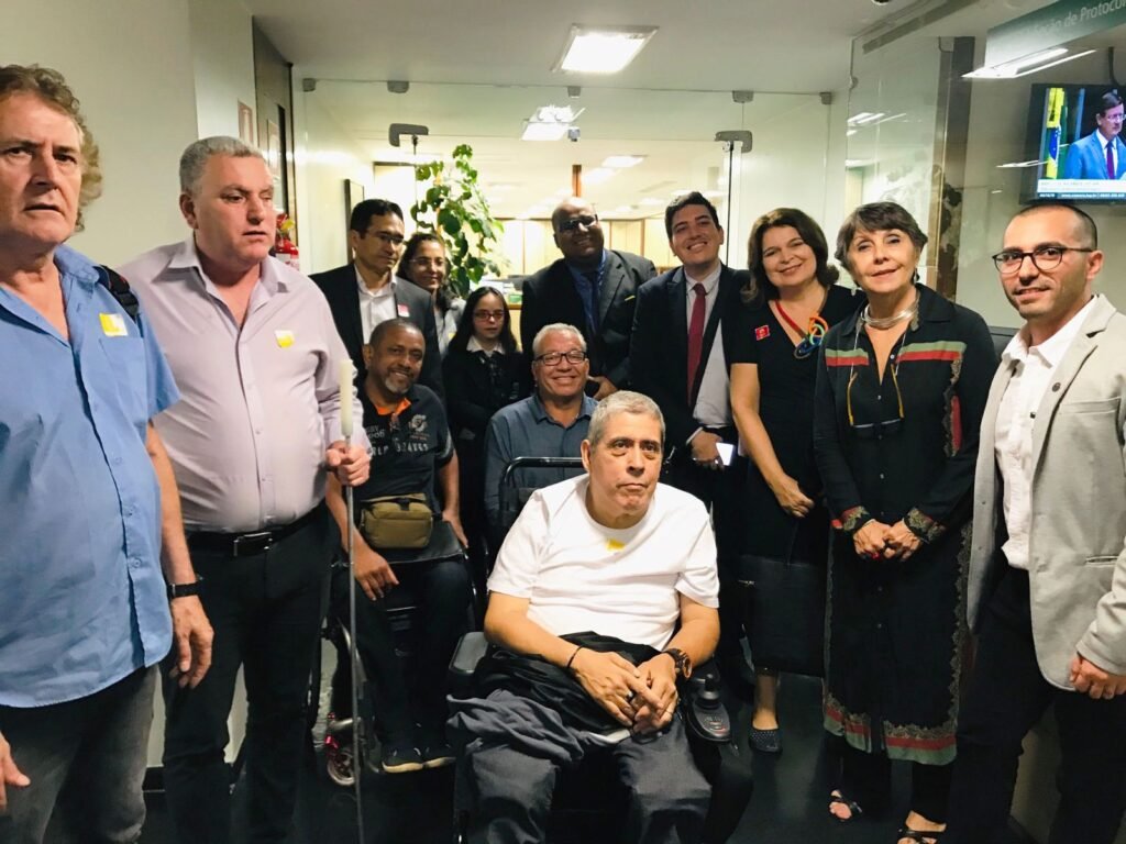 grupo de pessoas com e sem deficiência, entre elas 3 em cadeira de rodas, cego, com síndrome de Down, Patricia Almeida, Eduardo Cardoso e os Deputados Erika Kokay e Helder Salomão, num corredor da Câmara dos Deputados após protocolarem o PL 6256, em 3/12/19.
