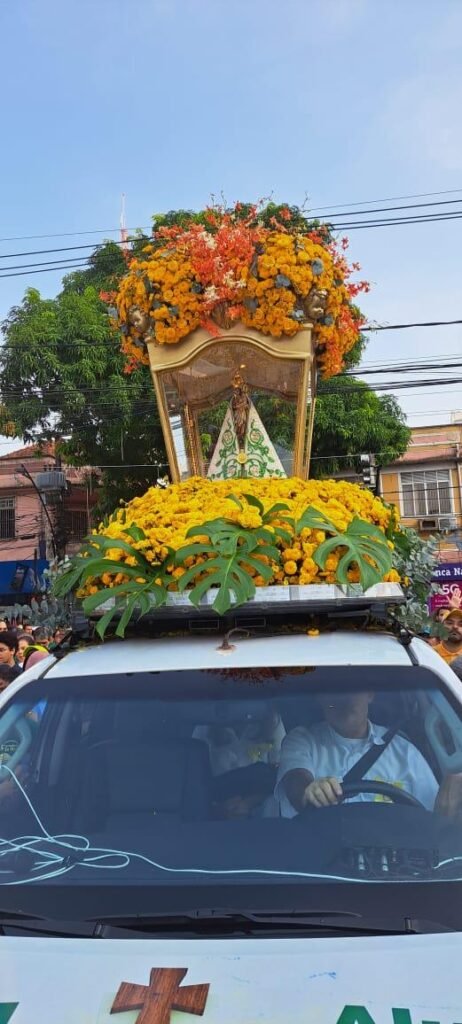 Um carro transporta a imagem de Nossa Senhora de Nazaré em uma berlinda ornamentada com muitas flores amarelas e laranjas. A pequena imagem da santa, vestida com manto branco e verde, está protegida por uma redoma de vidro. Ao redor, pessoas acompanham a procissão pelas ruas, sob um céu azul.
