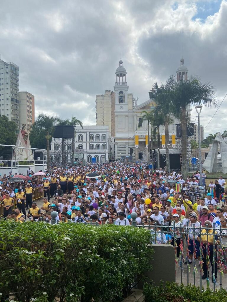 Uma multidão de fiéis se reúne em frente ao mesmo Santuário de Nossa Senhora de Nazaré, em Belém. O espaço está tomado por pessoas, muitas com camisetas brancas, bonés azuis e outros adereços. No fundo, a fachada da igreja aparece entre prédios e palmeiras, com bandeiras amarelas e brancas na entrada. O céu está nublado, mas a cena é vibrante pela grande quantidade de pessoas presentes na romaria