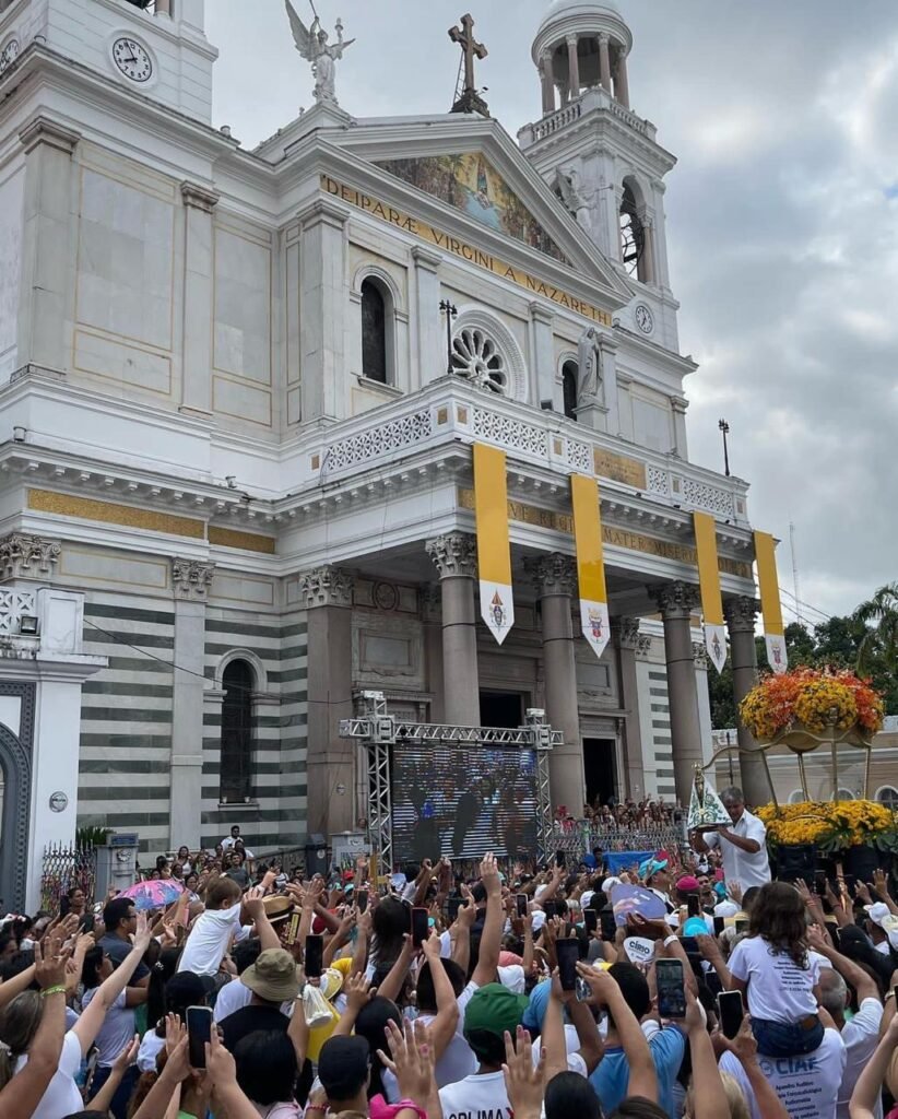 Uma multidão de fiéis se reúne em frente ao mesmo Santuário de Nossa Senhora de Nazaré, em Belém. O espaço está tomado por pessoas, muitas com camisetas brancas, bonés azuis e outros adereços. No fundo, a fachada da igreja aparece entre prédios e palmeiras, com bandeiras amarelas e brancas na entrada. O céu está nublado, mas a cena é vibrante pela grande quantidade de pessoas presentes na romaria.