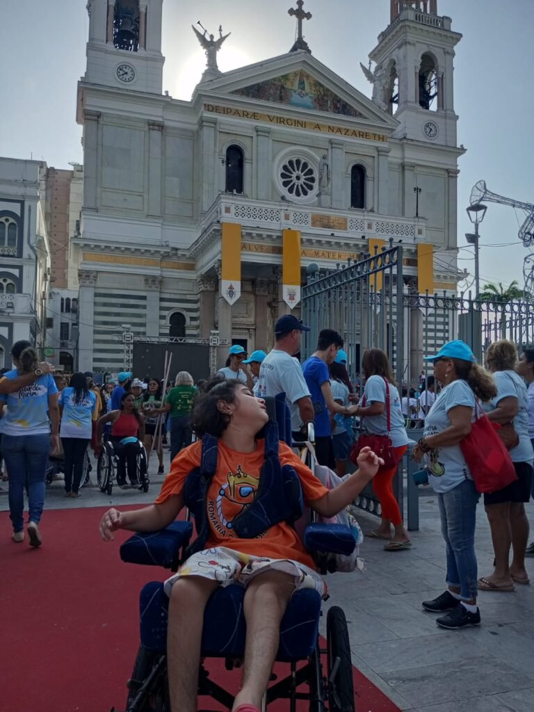 Uma pessoa em cadeira de rodas está em destaque no primeiro plano, vestindo camiseta laranja com estampa religiosa e shorts claros. Ao fundo, várias pessoas participam de um evento em frente a uma grande igreja, identificada como o Santuário de Nossa Senhora de Nazaré, em Belém do Pará. A fachada branca da igreja, com torres e relógios, aparece iluminada pelo sol que está atrás da construção. Há um tapete vermelho no chão e muitas pessoas usando camisetas e bonés coloridos.