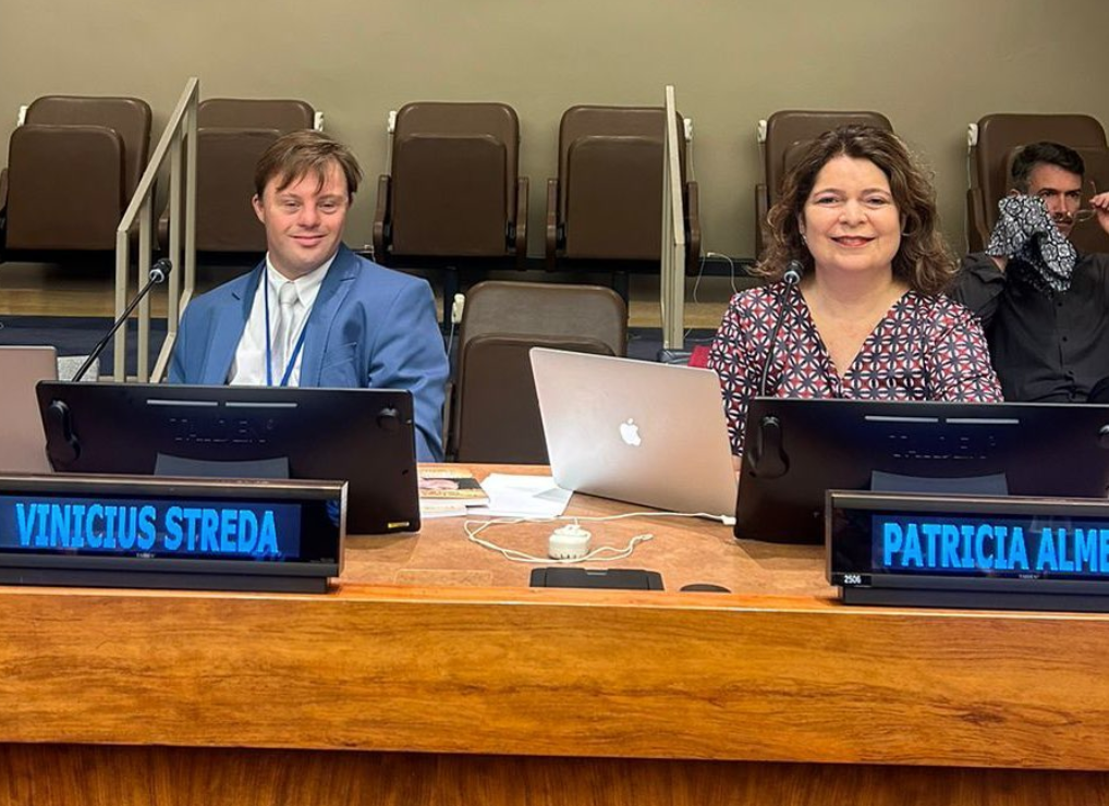 vinicius streda e patricia almeida sentados na mesa de debates da onu com seus nomes escritos no painel eletronico.