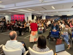 Uma mulher de cabelos brancos fala em pé, de costas para a câmera, em uma sala cheia de pessoas sentadas. Ela veste uma saia longa colorida e uma blusa vermelha. O público, diverso e atento, ocupa quase todas as cadeiras. No fundo, há um painel com letras grandes coloridas e uma câmera sobre tripé gravando o evento.