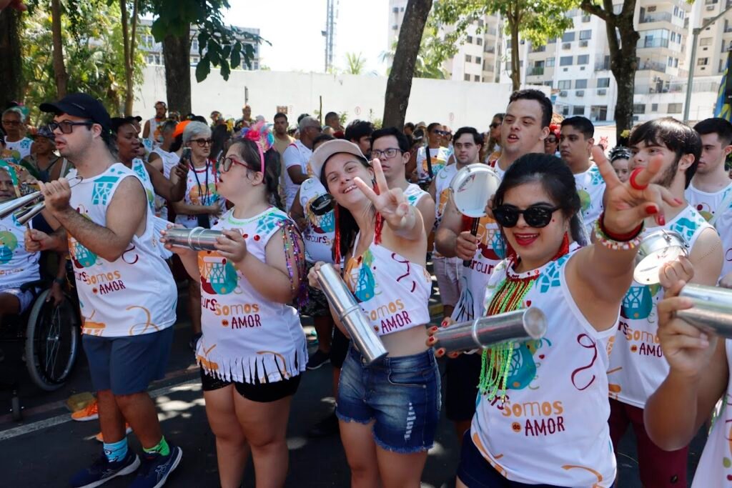 Grupo grande de pessoas em um desfile de rua. Elas usam camisetas brancas iguais e tocam instrumentos de percussão. Estão caminhando juntas, sorrindo e participando de um bloco de carnaval inclusivo.