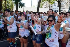 Grupo grande de pessoas em um desfile de rua. Elas usam camisetas brancas iguais e tocam instrumentos de percussão. Estão caminhando juntas, sorrindo e participando de um bloco de carnaval inclusivo.