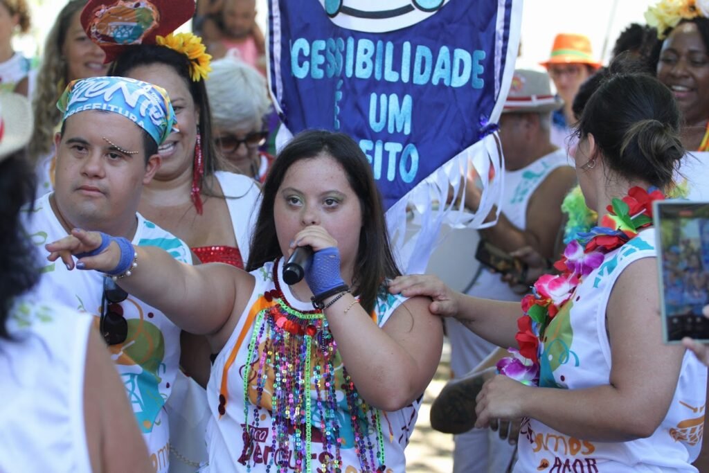 Pessoa com síndrome de Down segura um microfone e canta em meio ao bloco. Ao fundo, há uma faixa escrita “Acessibilidade é um direito”. Outras pessoas estão próximas, apoiando e participando.