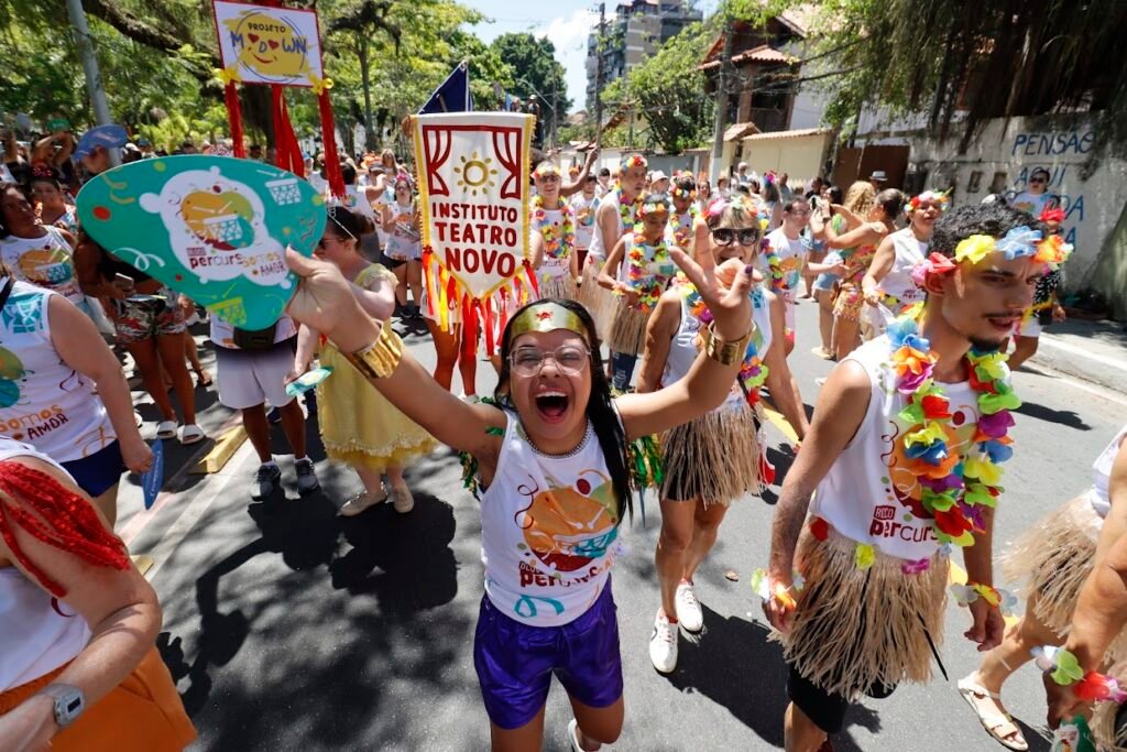 Pessoas do bloco caminham pela rua usando fantasias coloridas, colares e adereços. Uma pessoa no centro levanta os braços, sorrindo, demonstrando alegria e entusiasmo.