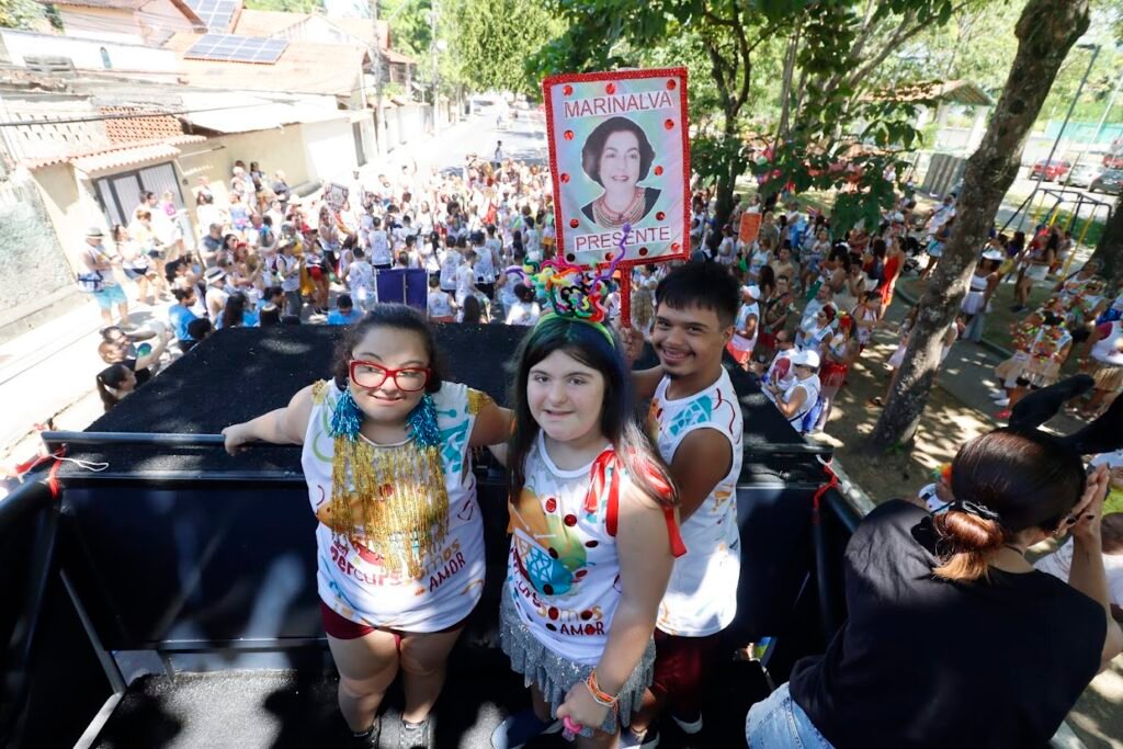Pessoas do bloco caminham juntas em uma rua arborizada. Há fantasias, música e dança. O ambiente é festivo, inclusivo e cheio de movimento.