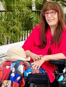 Rosangela Berman Bieler, mulher sorri com blusa vermelha, saia colorida, em cadeira de rodas.