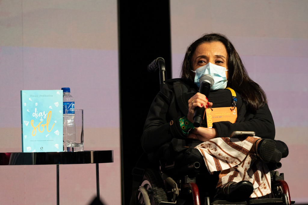 Thais Pessanha, uma mulher de longos cabelos escuros e usando uma máscara cirúrgica azul-clara, está sentada em sua cadeira de rodas em um palco, com o corpo ligeiramente voltado para a esquerda e olhando para o lado[cite: 5]. Ela veste um casaco preto fechado, calças escuras e sapatos, e segura um microfone com a mão direita próximo ao rosto, sugerindo que está falando[cite: 5]. Um crachá de identificação laranja e branco com o nome "THAIS PESSANHA" e a descrição "AGILE MASTER / SCRUM MASTER PETROBRAS" é visível em seu pescoço[cite: 5]. À sua esquerda, sobre uma pequena mesa redonda, está um livro com a capa azul e o título "dias de sol" em letras grandes e coloridas, com uma ilustração de sol e pássaros[cite: 5]. O fundo é um painel liso em tons de cinza e azul claro, com iluminação uniforme[cite: 5].