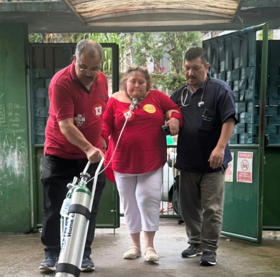 mulher com oxigenio e dois homens apoiando ao lado indo votar com camisa vermelha.
